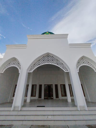 The image captures the facade of a white mosque with a green dome visible against a bright blue sky with wispy white clouds. The mosque features prominent arched entrances adorned with intricate geometric patterns. Several tall, narrow windows with dark frames are visible along the facade. A set of concrete steps leads up to the building. The foreground shows the pavement and the base of the steps, with a hint of foliage or ground cover. The location is identified as Keureuweungkrueng, Indonesia. No people are visible in the image.