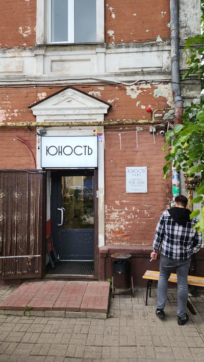 The image shows the entrance to the "Youth" cafe in Smolensk, Russia. The cafe's sign is displayed in Russian Cyrillic above the entrance. A second sign on the wall indicates the cafe's operating hours as 11:00 to 23:00, with no days off. A man in a checkered shirt and jeans is standing on the sidewalk next to a bench, holding a cigarette. The building is brick with peeling paint, suggesting its age. A window is visible to the left of the entrance. A section of brick pavement is to the right.