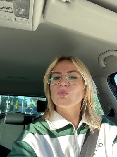 A blonde female is depicted from the mid-chest up, seated in what appears to be the front passenger or driver's seat of a vehicle. She wears square-framed, light-colored metallic glasses and has a "kissing" expression with pursed lips. Her attire consists of a green and white color-blocked collared sweatshirt or jacket with a visible zipper and a faint gold necklace. A dark seatbelt is secured across her chest. The vehicle's light gray interior, including an overhead console with lights, a sun visor, and a grab handle, is visible. Through the rear window, an exterior street scene is discernible, featuring a dark sedan, other vehicles, a barrier or fence, and several small, manicured trees under daylight conditions. The scene is located in the city of Antibes, France.