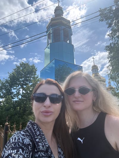 In a sunlit outdoor scene, two women are photographed in front of a striking modern church building. The church features a tall, multi-sided tower with a blue glass facade and a golden dome topped with a cross. To the right of the tower, another golden dome and a cross are visible, suggesting a larger complex. The sky above is a bright blue with scattered white clouds, and several power lines cross the frame. Lush green trees frame the right side of the image. In the foreground, the two women are positioned to capture a selfie. The woman on the left has brown hair and wears dark sunglasses and a black and white patterned shirt. The woman on the right has blonde hair, wears cat-eye sunglasses, and a black tank top with a white Puma logo. A decorative metal fence is visible to the left of the women. The scene appears to be set in Obukhivka, Ukraine, possibly near the Church of the Nativity of the Most Holy Mother of God, known for its unique modern architecture.