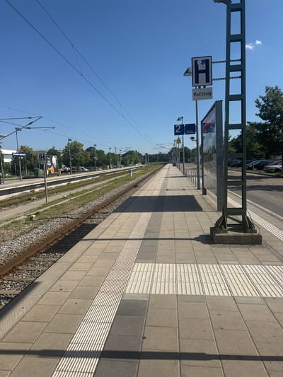 This is a vertical, eye-level shot of a train platform in Höhenkirchen-Siegertsbrunn, Germany, under a clear blue sky, suggesting a sunny day. The platform is paved with light grey and darker grey square tiles, with a textured strip of smaller tiles running along the edge closest to the tracks. The train tracks themselves are visible to the left of the platform, with ballast and overhead power lines.

A metal pole supports several signs. One sign is a blue rectangle with a white "H" inside a box, indicating a stop. Next to it is a white rectangular sign with "Kurzzug" written in black, meaning "short-haul train". Below these, a blue sign with a white "2" indicates platform number 2, and another blue sign says "Richtung Kreuzstraße", meaning "Direction Kreuzstraße". Further down the platform, another sign with a "2" is visible.

Other objects on the platform include a trash can and a large advertising display. In the background, beyond the tracks, there are trees and some parked cars. A shadow of a lamp post stretches across the platform tiles, suggesting the time of day is likely morning or afternoon. The scene conveys a sense of quiet anticipation, typical of a train station on a pleasant day. No people are visible in the frame.