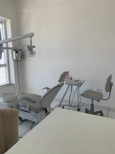 The image depicts the interior of an unoccupied dental examination room in Lauro de Freitas, Brazil. The room features white walls and a white tiled floor. A light gray, reclinable dental chair is positioned centrally, facing a large window on the left wall, which admits natural light. Attached to the dental chair unit on its left is a white ceramic spittoon. Overhead, an adjustable dental light with an articulation arm extends from the wall. To the right of the dental chair, a mobile dental unit equipped with various instruments and bottles stands. Adjacent to this unit, a gray, wheeled stool with a backrest is present, likely for a dentist or assistant. In the immediate foreground, the edge of a large, light-colored table is visible. A portion of a beige office chair is also visible in the lower-left foreground. No individuals are present, and no active procedures are occurring.