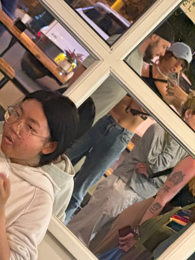 A fragmented view through a multi-pane window reflects an outdoor scene onto an interior setting where a person wearing glasses and a light-colored hoodie is visible. Outside, visible through the window, are various individuals, including one wearing a baseball cap and another with tattoos on their arm. The reflection shows a car or van, and a table with flowers. Based on the provided location of Sa Pa Ward, Vietnam, the scene is likely within a restaurant or establishment featuring a window. The angle suggests the photo was taken indoors, capturing both the interior subject and the reflected street activity.