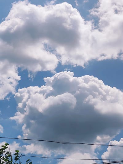The image shows a bright blue sky filled with large, fluffy white clouds. In the foreground, there are two dark horizontal wires stretching across the frame, with a few green leaves from a tree visible at the bottom left corner. The lighting suggests it is daytime, likely sunny. This scene is consistent with the typical sky and overhead infrastructure found in Durlești, Moldova, especially in residential or semi-rural areas where trees and utility lines are common. There are no people or specific actions depicted.