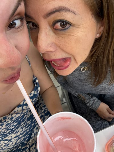 Two women take a selfie, their faces close together. The woman on top sticks her tongue out. A white cup containing a pink drink with a straw sits to the right of the women. The woman on the bottom is drinking from the straw. The woman on top wears a gray top and has a gold earring. The woman on the bottom wears a blue patterned dress.