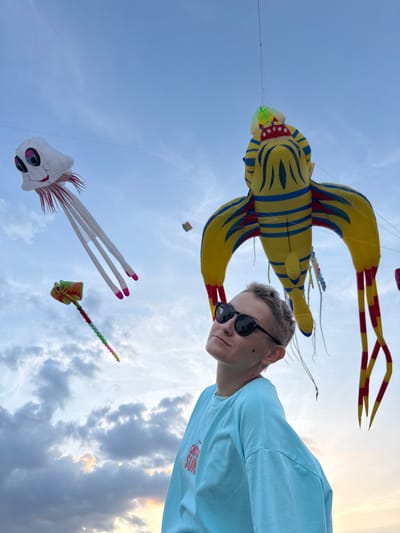 A person with short, blonde hair and sunglasses is looking up at the sky. They are wearing a light blue t-shirt with red text that reads "CATCH SUN." In the background, several kites are flying against a cloudy sky. One large kite is yellow with blue stripes and resembles a fish or sea creature. Another kite is white with a skull-like face and pink-tipped tentacles. A smaller, colorful kite with a striped tail is also visible. The time of day appears to be late afternoon or early evening, as suggested by the warm glow of the setting sun peeking through the clouds. The scene suggests an outdoor activity, likely a kite festival or a leisurely day of kite flying, taking place in Quy Nhon Ward, Vietnam. The overall mood seems relaxed and enjoyable.