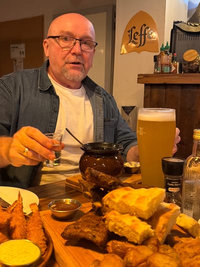 A bald man wearing glasses and a denim shirt sits at a table with a large beer glass, a ceramic pot with a long spout, and a shot glass in his hand. A wooden board displays bread, fried food, and two small bowls of dipping sauce. Shelves in the background hold various bottles, including hot sauce and spices. A "Leffe" sign hangs on the wall.