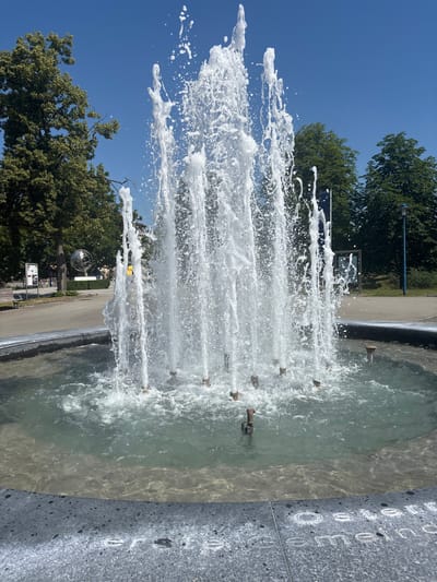 This media file captures a dynamic water fountain in a public outdoor setting on a clear, sunny day. The primary subject is the fountain's water jets, which are spraying upwards and outwards, creating a dynamic, frothy display against a bright blue sky and lush green trees.
The fountain appears to be part of a larger structure, with a textured, dark gray stone border visible on the right side, inscribed with white lettering that is partially obscured and difficult to read clearly. Several small, metal spouts are visible protruding from the fountain's base, from which the water jets originate.
In the background, trees line a path or walkway, suggesting a park or urban green space. A spherical, metallic sculpture is visible in the distance, adding an artistic element to the scene. There are also signs and structures that hint at a public area, possibly a plaza or promenade. The time of day is clearly daytime, indicated by the bright sunlight and clear sky. The weather is pleasant and sunny.
No people are clearly visible or identifiable within the frame, focusing the attention solely on the fountain and its surroundings. The overall emotion evoked is one of freshness, energy, and a bright, lively atmosphere. The location context places this scene in Gebersdorf, Austria.