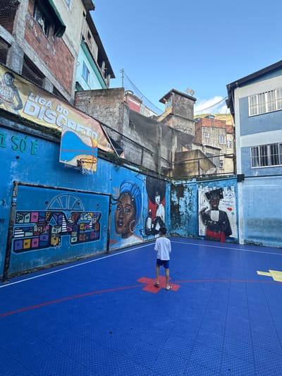 This image depicts an outdoor sports court located within a favela in Rio de Janeiro, Brazil. The court features a bright blue modular tiled surface with red and white demarcation lines, and a red and yellow cross-shaped marking. A single male child, dressed in a white t-shirt, blue shorts, and flip-flops, stands with his back to the viewer in the foreground, facing the court's perimeter wall.
The perimeter walls are painted blue and adorned with extensive murals. On the left wall, a mural displays colorful, geometric representations of stacked houses, adjacent to a large portrait of a woman with dark skin, wearing a blue headwrap and jewelry. Above this section, a basketball hoop with a custom-painted backboard, depicting a landscape, is affixed to the wall. Above the hoop, a yellow banner stretched across the upper wall reads "LIGA DO DISCRETO" and features an illustration of a basketball player. The right wall contains multiple large murals, including a portrait of a person with an afro hairstyle and boxing gloves (partially obscured by the child), and another portrait of a young person in a black graduation cap and gown, holding a microphone, with text inscribed alongside.
Surrounding the court, multi-story residential buildings, constructed from exposed brick and concrete, rise irregularly, characteristic of favela development. These buildings are painted in various tones including grey, blue, and light green, featuring numerous windows, some with metal grates. Black netting is stretched above and between several structures, likely for safety or as a ball barrier. A clear blue sky is visible overhead, with distant hills and additional buildings, including rooftop water tanks, discernable on higher ground.
