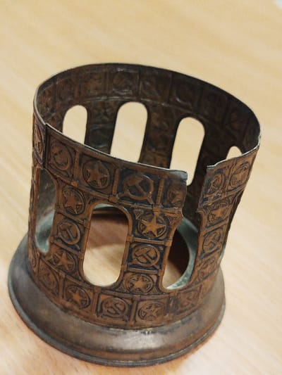 The image shows a cylindrical metal cup holder with a decorative design of Soviet symbols, including stars and the hammer and sickle. The cup holder has an open design with vertical slots. The metal appears to be aged and has a dark brown or bronze finish. It is placed on a light-colored wooden surface. It's highly unlikely, based on the image alone, to associate it with a specific location like the city of Боровково, Russia. More information would be needed to establish a location.