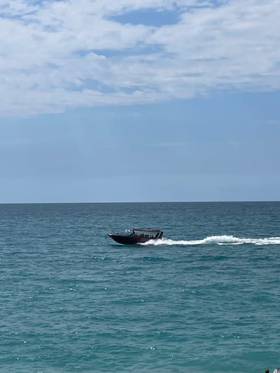 A speed boat is traversing the turquoise waters of the Black Sea, leaving a white wake behind it. The boat appears to be carrying several passengers. The water is calm with small ripples. In the background, the horizon is visible, with a clear blue sky dotted with white clouds. The scene is likely from the city of Gagra, Abkhazia, a popular Black Sea resort known for its coastline and recreational activities.