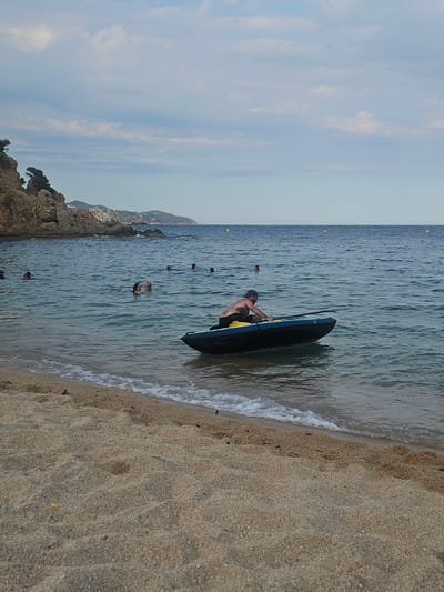 The image depicts a beach scene in Blanes, Spain. The primary subject is a person in a black kayak, positioned at the edge of the water where it meets the sand. The person is wearing a dark cap and yellow shorts, and appears to be preparing to enter or exit the kayak. The water is calm with gentle waves lapping at the shore. In the background, several other people are visible swimming or wading in the sea. Rocky outcrops and lush vegetation line the coastline in the distance, suggesting a scenic, natural environment. The sky is mostly clear with some wispy clouds, indicating fair weather, likely during the daytime. The overall mood is relaxed and recreational, as people enjoy a day at the beach. The sandy texture of the beach and the clear blue water are prominent details. No visible text is present in the image.