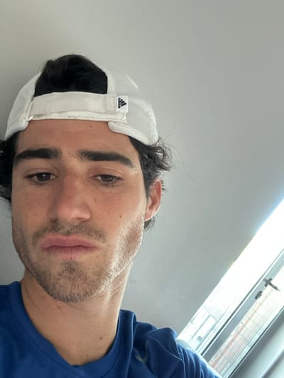 A male individual is depicted in a close-up, chest-up portrait taken indoors. He wears a white Adidas baseball cap reversed, with the Adidas logo visible on the back strap, and a blue short-sleeved shirt. His dark hair is visible, and he has light facial hair. His expression is neutral, with his eyes directed slightly downward. The background consists of a light-colored wall or ceiling. A portion of a window frame, likely a skylight or attic window, is visible in the lower right, admitting bright natural light. No specific landmarks or distinguishing features are present to determine a precise location within Giebułtów, Poland; the scene is consistent with a residential interior.