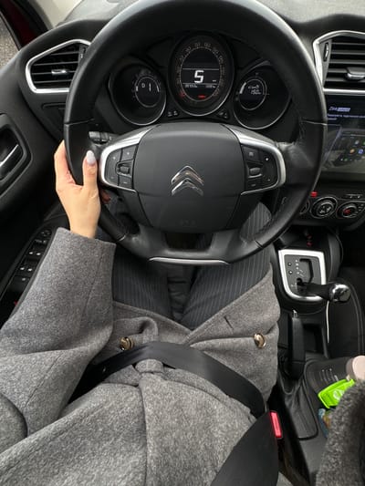 The image shows the interior of a Citroen car. A person wearing a gray coat and striped pants is seated in the driver's seat, with the seatbelt fastened. Their left hand is on the steering wheel. The dashboard is visible, displaying a speed of 5 km/h, and the car is in Drive ("D"). The gear shift is in the automatic position. A drink container is in the cup holder. The location is unknown. The presence of Russian text on the infotainment display suggests the car is in Russia but there is no indication that it is in Dubki (Gp Kubinka).