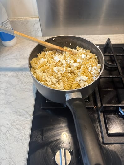 A top-down shot shows a black saucepan filled with cooked fusilli pasta and chunks of white cheese. A wooden spatula rests inside the pan. The saucepan sits on a black glass stovetop. The stovetop is situated on a countertop featuring a white marble-like pattern. A portion of a white plastic bottle with a blue cap is visible in the lower left corner. The setting is likely a domestic kitchen in Swindon, United Kingdom. The focus is on the food preparation. No people are visible.