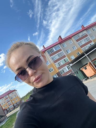 A woman with blond hair and sunglasses is taking a selfie outdoors. In the background, there are multi-story buildings with yellow, gray, and red trim, suggesting an urban or suburban setting in Высокий, Russia. The sky is blue with some clouds.