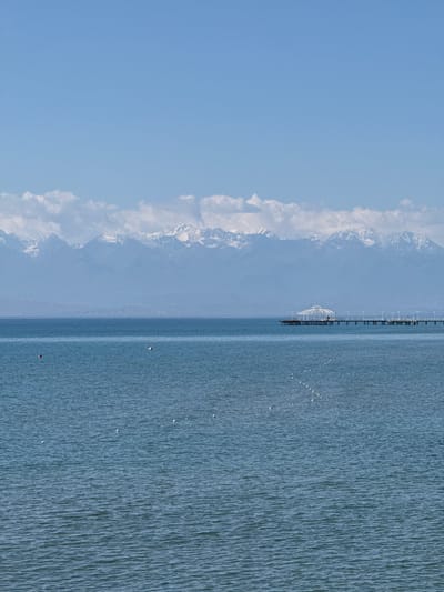 The image depicts a serene seascape with a distant, snow-capped mountain range under a clear blue sky. In the foreground, the calm blue water stretches to the horizon, with a series of white buoys forming a subtle line across the surface. To the right, a long pier extends into the water, featuring a white, open-air structure at its end, resembling a gazebo. The pier is lined with lights. On the water, closer to the shore, there are two buoys, one red and one white. The scene appears to be a tranquil day at a coastal or lakeside location, possibly in Koshkol', Kyrgyzstan, given the geographical context provided, with the mountains suggesting a proximity to a significant mountain range like the Tian Shan.