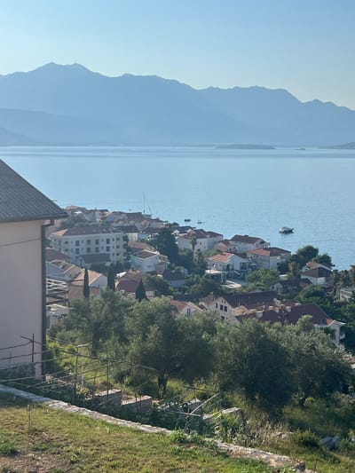 In Đenovići, Montenegro, a panoramic view captures a tranquil coastal scene under a clear sky. The foreground is dominated by a grassy slope with a stone wall and a rustic trellised structure, suggesting a garden or vineyard. Beyond this, a cluster of houses with red-tiled roofs nestles among lush green trees, indicative of a Mediterranean landscape. The buildings are of varying sizes and architectural styles, with some featuring multiple stories and balconies.
In the mid-ground, a body of calm, blue water stretches out, dotted with a few boats, including a sailboat and a motorboat. The water reflects the bright sky, creating a serene atmosphere.
In the background, a series of majestic mountains rise, their peaks softened by a light haze. The largest mountain in the distance has a visible structure at its summit, possibly a telecommunications tower. The overall impression is one of a peaceful and scenic coastal settlement.