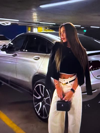 A woman with long, dark hair stands in a dimly lit parking garage, posing next to a silver car. She is wearing a black crop top, white pants, and a black jacket draped over her shoulders. She is holding a small black purse in her right hand. The garage has a concrete ceiling with fluorescent lighting and a blue and yellow painted wall in the background. A yellow line is visible on the floor, indicating a parking space. This scene is located in the city of Tomai, Moldova.
