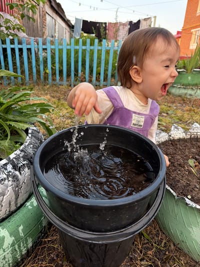 In Vikulovo, Russia, a toddler, wearing a light-colored long-sleeved shirt and purple overalls, joyfully splashes water in a black bucket with their right hand. The child's mouth is wide open, and they appear to be laughing. The bucket is filled with murky water, and splashes are visible in the air. The scene unfolds in an outdoor setting, likely a backyard, with a wooden fence painted blue in the background. Clothes hang on a clothesline in the distance, and structures of a house are visible. The ground is covered with dry grass and soil, with some greenery and tires repurposed as planters in the foreground. The overall ambiance suggests a warm, informal day spent outdoors.