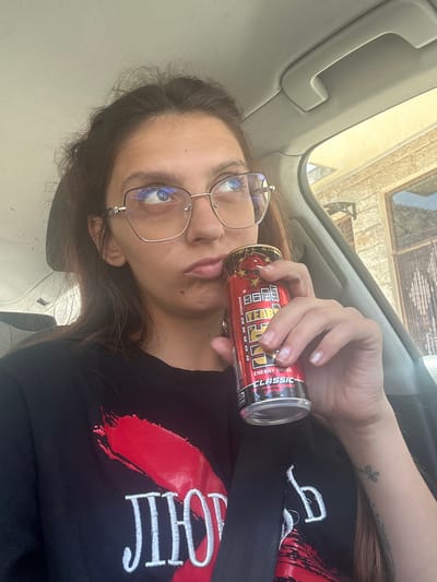 A young woman with brown hair and glasses is sitting in the driver's seat of a car, holding a can of "Hell Energy Drink Classic 30th Anniversary Edition." She is wearing a black t-shirt with a red graphic and white text. The car appears to be parked on a street in the city of Tryavna, Bulgaria, as indicated by the architecture visible through the car window. The background shows a building with light-colored stone walls and windows. The overall scene is a candid shot, likely a selfie, taken during the daytime.