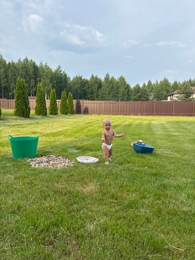 In a suburban backyard in Борисьево Днп, Russia, a young child wearing a diaper stands on a manicured green lawn. The child is facing the camera with their arms outstretched. To the left of the child, a large green bucket sits on the grass next to a pile of grey pebbles and a white plate with a pink object. Further to the right, a blue plastic tub is placed on the lawn. In the background, a brown wooden fence encloses the yard, beyond which a dense line of trees forms a natural barrier. A building with a brown roof is visible in the far distance, peeking out from behind the trees. The sky is cloudy with patches of blue.