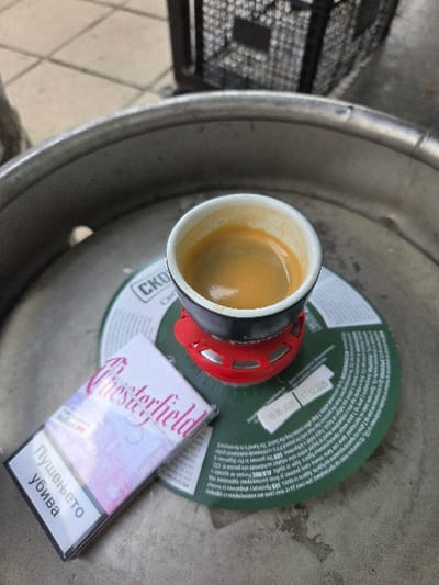 The image displays a close-up, elevated view of an espresso cup and a pack of cigarettes positioned on the metallic top of a beverage keg, set within the city of Gevgelija, North Macedonia. The small white ceramic cup, dark on the exterior, contains espresso topped with a layer of crema, and rests on a red plastic multi-faceted base. Beneath the cup and base is a circular dark green coaster featuring Cyrillic text, including "СКО" and dates "15.06.2023" and "17.12.2025." To the left of the cup, a white and pink pack of Chesterfield cigarettes is visible, bearing the health warning "Пушењето убива" (Smoking kills) in Cyrillic. The metallic surface of the keg exhibits wear and minor indentations. In the blurred background, tiled flooring is partially visible to the upper left, and a dark mesh-like object, possibly a crate or basket, is present to the upper right. No individuals are visible, and no active actions are depicted.