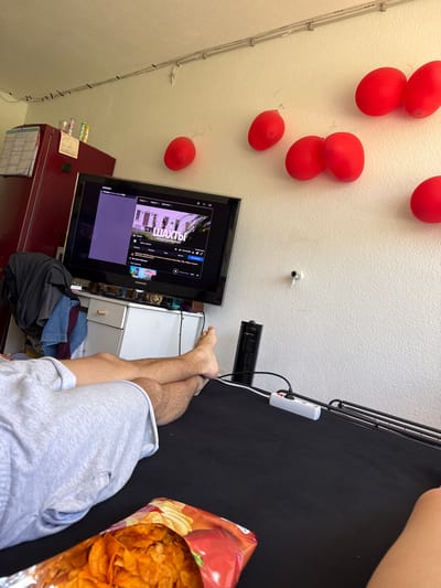 The image depicts an indoor scene, likely a living room. In the foreground, a person's legs are visible, clad in light gray shorts, resting on a black surface, possibly a bed or sofa. Next to their legs is an open bag of potato chips. In the midground, a television screen displays a streaming service with a title that appears to be in Russian, showing a scene from a show titled "ШАХТЫ". Below the television, a white cabinet with drawers is visible, and a dark grey t-shirt is draped over the left side of the cabinet. The background features a white wall adorned with several red balloons. A red refrigerator is positioned to the left of the television. A power strip with multiple outlets and connected cables is visible on the black surface in the midground. The overall impression is one of casual relaxation and entertainment. There are no specific visual cues to indicate the city of Lörrach, Germany.