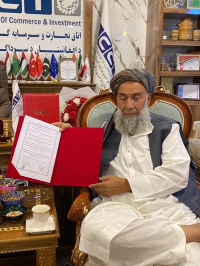 In an indoor setting, a man with a long white beard and a turban, possibly an Afghan official, is seated in an ornate chair. He is holding a red folder with a document inside. Behind him are flags, including those of Afghanistan, China, Pakistan, and the European Union, along with a sign for the Afghan Chamber of Commerce & Investment. Additional details in the background include shelves with decorative items, indicating an official or formal setting. No specific evidence indicates this to be Arghandab, Afghanistan.