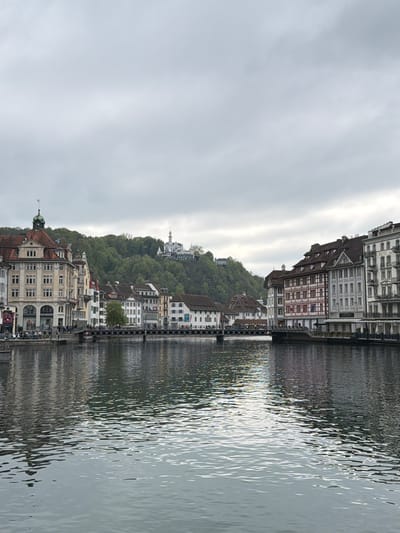 A cityscape on the water in Lucerne, Switzerland. Buildings line the canal, with a hill in the background topped by a distinctive white building. A bridge spans the water in the middle distance. The water reflects the buildings and sky. The sky is overcast.