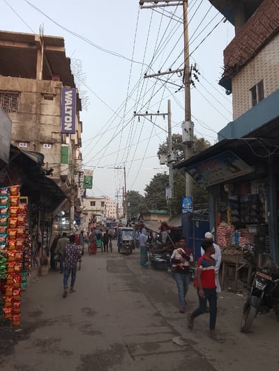 The image depicts a ground-level view of a narrow, urban street in Tarabo, Bangladesh, under an pale, undifferentiated sky. A dense network of black utility wires is strung overhead, connecting to multiple concrete utility poles, two of which are equipped with gray transformers.
Buildings of varying heights and construction materials line both sides of the street. On the left, a multi-story concrete structure displays a prominent vertical blue sign with the text "WALTON" in white. Below this, an open-air display rack holds numerous brightly colored packaged snack items. On the right, a building with a blue upper section and reddish brick features a ground-level shop under a dark awning, where sacks of produce and other goods are visible. A black motorcycle is parked on the far right.
The street surface is a worn, gray asphalt or concrete with visible cracks and patches. Pedestrian traffic is present. In the foreground right, a male subject wearing a red and blue striped shirt walks towards the left side of the frame, while another male subject in a white tunic and head covering stands near the shop stall. Further left, a male subject carries a small child. In the mid-ground, a blue auto-rickshaw and a red bicycle rickshaw are positioned further down the street, both facing away from the viewer. Several other individuals are dispersed throughout the scene, walking or standing. Utility poles feature attached banners and signs with Bengali text, including a blue banner with a portrait of a male figure.