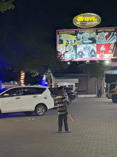 The image depicts an outdoor scene at night in Keudeteupinpunti, Indonesia. A large illuminated billboard dominates the upper portion of the frame, advertising "DYL DAYAH LIVING," which appears to be a department store or shopping complex. The billboard is divided into six sections showcasing various products and services, including "HOUSEWARE," "FURNITURE," "KARPET-PERMADANI" (Carpets/Rugs), "MADINAH ROPIE REST AREA," "BOISS BOUTIQUE," and "D'FRIED CHICKEN."
In the foreground, a white minivan is parked on a patterned brick surface. A man wearing a striped shirt and dark pants stands near the vehicle, holding a red baton. To the right of the minivan, a portion of a yellow truck is visible. In the background, several trees with decorative lights are present, along with a brick building and a ladder leaning against it. A person is seated on the ground near the ladder. The overall lighting suggests it is nighttime.