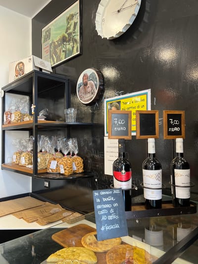 The image depicts the interior of a small shop, likely a bakery or a specialty food store, located in Bari, Italy. The shop has a rustic and slightly cluttered aesthetic, with a black, textured wall as the backdrop.
On the shelves are various food items packaged in clear plastic bags, appearing to be taralli, a traditional Italian snack. These are arranged on two black metal shelves. Above the shelves, on the wall, are several decorative items and displays. A framed movie poster, possibly from an Italian Western film, is prominently displayed. To its right, a circular decorative item with a picture of a man's face is visible, and above that, a large, white, bottle-cap-shaped clock with Roman numerals.
In the foreground, on a lower shelf, three bottles of wine are displayed on a dark metal rack. Each bottle has a label, and two of them are advertising "Salice Salentino." Above the wine bottles, three small wooden-framed chalkboards are mounted on the wall. Two of these chalkboards display prices: "7,00 EURO." The third chalkboard is blank.
To the left of the wine bottles and chalkboards, a sign written in chalk on a dark blue background states: "Da Gennaio 2022. le buste avranno un costo di 10 cents. #focaccia." This indicates that as of January 2022, bags would cost 10 cents, and mentions "focaccia," a type of Italian flatbread.
Below the shelves, on a white counter, are stacks of brown paper bags, some of which appear to have a logo or text printed on them, possibly related to "focaccia." Further down, in a glass display case, pieces of focaccia and other baked goods are visible. One piece of focaccia in the foreground is topped with what appear to be slices of lemon.
The overall impression is of a local establishment selling regional products. The presence of the movie poster and the specific mention of "focaccia" suggest a connection to Puglian culinary traditions. The lighting is somewhat dim, with spotlights highlighting specific areas of the display.