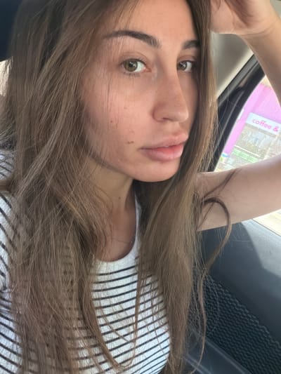 A close-up, selfie-style photograph shows a young woman with long, brown hair, wearing a white and black striped t-shirt. Her left hand is resting on her head. She is seated in the passenger seat of a car, as indicated by the car door and window visible in the frame. Through the car window, a pink building with the text "coffee &" is visible, suggesting a commercial establishment. The lighting suggests it is daytime. The specific location in Larnaca, Cyprus, cannot be determined from the visual information provided.