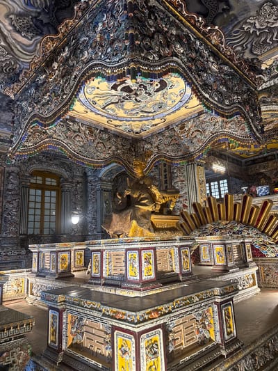 The image shows the ornate ceiling of a building, likely a temple or palace, in Thủy Xuân Ward, Vietnam. The ceiling is elaborately decorated with intricate carvings, paintings, and mosaics. The artwork depicts various figures and scenes, possibly from Buddhist or Taoist mythology. A golden statue is visible, suggesting the religious significance of the building. The architectural style is traditional Vietnamese, with a blend of Chinese and local influences. Windows provide natural light, illuminating the detailed artwork.