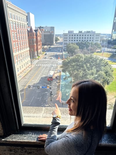 A woman with long brown hair leans out a window, pointing down with her right hand. A gold wristwatch is visible on her wrist. Outside the window, a cityscape is visible, including tall buildings, trees, and a street with traffic lights. Her reflection can be seen in the window glass. The window frame is dark, and the wall around it appears textured.