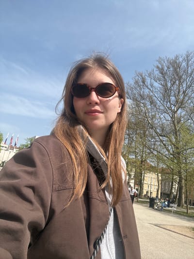 A young woman poses for a selfie in Ljubljana, Slovenia. She is wearing sunglasses and a brown jacket, with a gray hooded sweatshirt underneath. Her hair is long and brown. Behind her, there is a clear blue sky with some trees and buildings. Flags are visible in the background. In the distance, people are sitting on benches in a park-like setting. The ground is paved.
