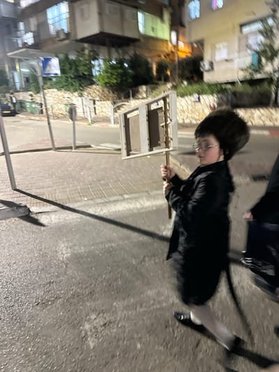 The image captures a nighttime street scene in Bnei Brak, Israel. A young boy, approximately 6-10 years old, stands on a cobblestone-patterned sidewalk in the foreground, facing right in profile. He wears a black fur hat, a dark long coat, light-colored stockings, and dark shoes, along with round-rimmed glasses. The boy holds a white-framed object upright with both hands on a wooden pole. This object, resembling a large, simple wooden frame, contains two rectangular wooden panels and features small, blurred decorative elements or lights on its upper right side. To the boy's right, a second individual is partially visible, primarily their lower torso and legs, also in dark attire and appearing to be in motion. The foreground consists of a dark asphalt street. In the mid-ground and background, residential buildings with illuminated windows are visible. Other environmental details include a low stone wall with vegetation, a street pole with a sign, and partially visible green dumpsters. Artificial lighting illuminates the scene, casting shadows.