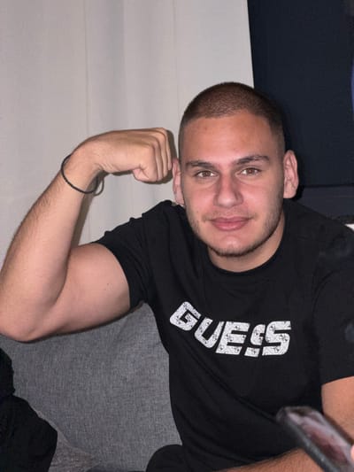 A young adult male with a buzzed haircut and a light beard is seated on a grey couch, with his left arm flexed to display his bicep. He is wearing a black t-shirt with the "GUESS" logo printed in white across the chest. A thin black bracelet is visible on his left wrist. The background consists of a light-colored wall with vertical lines, possibly a curtain or textured wallpaper, and a dark navy-blue surface on the right side of the frame. The overall setting appears to be an indoor environment, likely a residential space in Musachevo, Bulgaria.