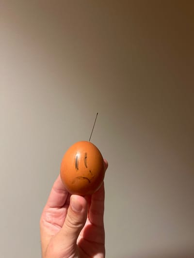 A light brown, ovoid egg with a sad face drawn in dark marker, featuring two vertical lines for eyes and an inverted U-shape for a mouth, is held by a light-skinned human hand. A thin, metallic needle is inserted vertically into the top of the egg, angled slightly upward and to the right. The hand grips the lower portion of the egg with the thumb and fingers. The background is a uniformly illuminated, plain wall of a light, neutral color. This scene is located in Aldoar, Foz do Douro e Nevogilde, Portugal.