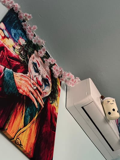 The image shows an indoor scene, with a portion of a wall and ceiling visible. On the wall, a large framed artwork depicting the character Joker is displayed. The artwork is adorned with artificial pink flowers that drape over its top edge. To the right of the artwork, a white air conditioning unit is mounted on the wall. A small, plush toy resembling a cup of bubble tea is attached to the air conditioning unit. The scene does not provide any information or context that would indicate a location in the city of Debelt, Bulgaria.
