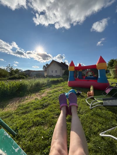 The image depicts a sunny outdoor scene in the village of Dunilovo, Russia. In the foreground, a person's legs, wearing purple patterned clogs, are extended towards the viewer. The ground is covered in lush green grass. To the right, a colorful inflatable bounce house is set up. Various items are scattered on the grass around the bounce house, including what appears to be a leaf blower, a blue bag, and parts of lawn chairs. In the background, a light-colored house with a prominent roofline stands. Beyond the house, there are trees and other vegetation, suggesting a rural or semi-rural setting. The sky is blue with scattered white clouds, and the sun is shining brightly, casting rays of light.