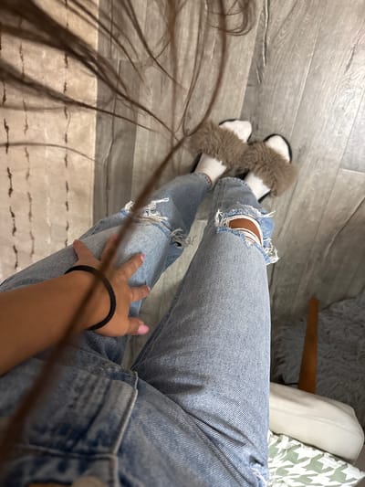The image shows a person's legs, wearing ripped blue jeans and white socks with fuzzy slippers. Their right hand, adorned with a black hair tie on the wrist and pink nail polish, rests on their thigh. Long, dark hair is draped across the scene, obscuring parts of the background. The floor appears to be light-colored wood planks. A blurry object, possibly a stool or chair, is visible in the lower right corner. The scene is indoors. The location of Basarabeasca, Moldova, is not evident from the visual information provided in the image.