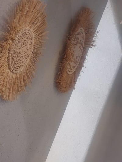 Two decorative wall hangings made of natural fibers and carved wood are affixed to a textured wall. The hangings are circular with a central carved design and a fringe of straw-like material. One hanging is positioned to the left of the image and is larger than the one on the right. The wall appears to be an interior surface, possibly a ceiling given the angle of the lighting. A diagonal shadow falls across the right side of the image, suggesting a light source from the upper left. The location is not specified in the image itself, but if it were in Kyzylorda, Kazakhstan, it would likely be an indoor decorative element within a residence or public space.