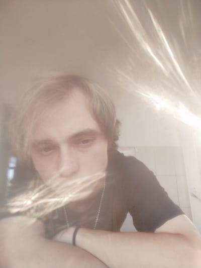 The image shows a young adult with light-colored hair looking down and to the right. They are wearing a dark t-shirt and a silver chain necklace. The subject's arms are crossed, resting on a surface that is not fully visible. The background appears to be indoors, with a tiled wall to the right. There are strong light refractions or lens flares present in the image, creating bright, elongated streaks of light across the scene, particularly in the upper right and lower left portions. The overall lighting is bright and somewhat diffused, with a warm tone. There are no discernible elements in the image that suggest a specific location within the city of Salsk, Russia.
