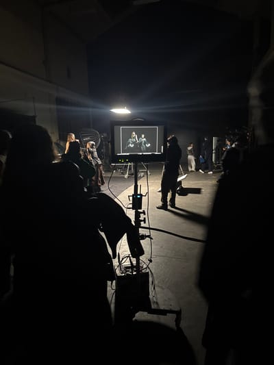 The image depicts an active photography or video production set located within a large, dimly lit industrial studio in Ivry-sur-Seine, France. A central, prominent monitor on a wheeled stand displays a high-contrast image of two individuals, one female with long hair and one male, both in dark attire, against a dark background, indicating a captured photograph or video frame.

In the mid-ground to the left of the monitor, a female with blonde hair, wearing a black top, is visible, possibly posing or being attended to by another individual in dark clothing. Behind them, a large, silver parabolic softbox and another light source provide illumination. To the right of the monitor, a male figure stands with his back to the viewer, observing the screen. Further into the background on the right, several individuals, some in lighter casual clothing, are standing or moving, appearing to be crew members or staff.

In the immediate foreground on the left, a partial silhouette of a person observes the scene. A stand with visible electronic equipment and multiple cables is positioned in front of them. The environment features a concrete floor, dark walls, and high ceilings, with what appears to be a large industrial door or partition visible in the background. The scene is predominantly dark, with key lighting focused on the primary subjects and the monitor, creating strong silhouettes of foreground and mid-ground figures.