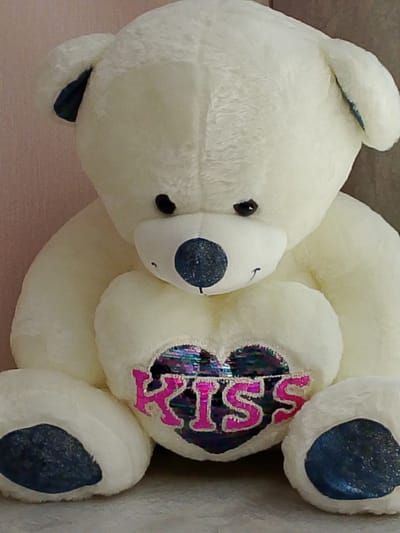 A close-up, head-on shot shows a large, cream-colored stuffed teddy bear sitting upright. The bear has dark eyes, a dark, sequined nose, and a small embroidered smile. On its chest, a heart-shaped patch is adorned with iridescent sequins and the word "KISS" spelled out in glittery pink letters. The bear's paws are visible, with dark blue, sparkly accents on the soles. The background is softly blurred, showing a pale pink textured surface to the left and a gray surface to the right. There is no indication of the city of Yanino-1, Russia in the image.