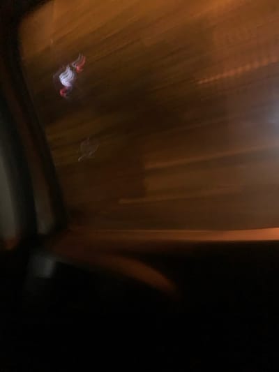 The image presents a severely motion-blurred nighttime scene, captured from inside a moving vehicle in Lauro De Freitas, Brazil. The foreground, occupying the lower half of the frame, is dark and indistinct, consistent with a vehicle's interior. The background, visible through what appears to be a window, consists of horizontal streaks of warm, orange-brown ambient light, indicative of streetlights or other urban illumination viewed during rapid transit. In the upper-left quadrant, two highly blurred and amorphous luminous anomalies are present. The upper anomaly appears as an indistinct swirl of white and red light. Directly below it, a second, lighter-toned, less defined amorphous shape is discernible. The extreme horizontal streaking across the entire external view indicates significant motion during exposure. No identifiable individuals or specific objects with clear forms are present due to the pervasive blur.