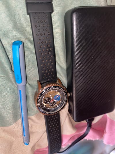 The image presents a still-life arrangement of personal items on a soft, textured fabric surface, likely indoors in Hayin Banki, Nigeria.
The main subjects include a black rectangular electronic device, a watch, and a pen. The black device, possibly a power bank or external hard drive, is positioned at the top of the frame. It features a textured surface with diagonal lines and exhibits visible scuff marks, indicating usage. A black cable is plugged into its right side.
Below the black device lies a watch with a distinctive appearance. Its casing appears to be bronze or rose gold, showing some signs of wear and patina. The watch face is a light brown or golden color, accented with blue and black sub-dials, characteristic of a chronograph. The black bezel around the dial has white numerical markings, with '50', '40', and '30' clearly visible. The watch is fitted with a black rubber strap, which is perforated with small circular patterns.
In the foreground, a gray and blue pen is visible. The pen has a light gray body with a bright blue clip and blue accents. The brand name "Flair" is visible on the blue clip. The items rest on a light green or teal-colored fabric, which appears to be a bedsheet or blanket. In the upper right background, a section of pink and white striped fabric is partially visible. The lighting is relatively dim, suggesting an indoor setting without strong direct light. No specific activity is taking place beyond the arrangement of these objects.