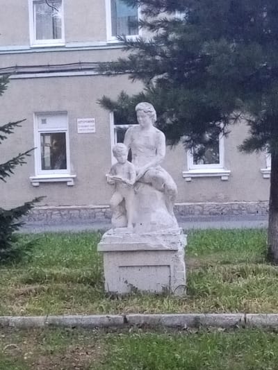 The image depicts a white stone sculpture of a woman and a child in front of a light-colored building with several windows. The sculpture is situated on a grey stone pedestal, which is placed on a patch of grass. A large evergreen tree is partially visible in the foreground and on the right side of the image. The building appears to be two stories tall, with white-trimmed windows. One of the windows on the first floor has a sign above it that reads "ОСТОРОЖНО" (Caution). The overall environment suggests an outdoor setting in a public space, likely a park or the grounds of an institution. The style of the sculpture and the signage hint at a location in Russia. Specifically, this sculpture is located in the city of Yurga, Russia, at the address Komsomolskaya Street, 17A, which is the site of the Yurga Branch of the Tomsk State Pedagogical University.